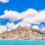 En ekstremt detaljeret udsigt over Eivissas gamle bymidte og marina, lys himmel, maleriske skyer, et fortøjet sejlskib, den ikoniske skyline i Dalt Vila domineret af katedralkirken Santa Maria de les Neus. Fremkaldt fra RAW.