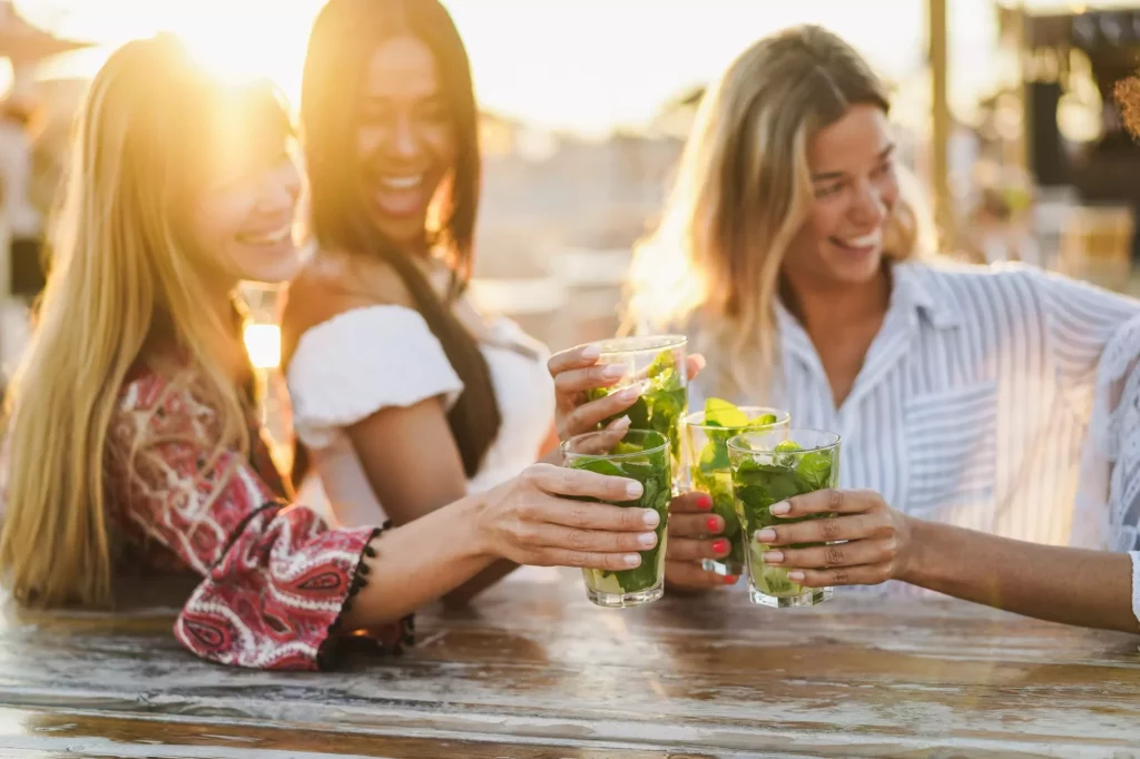 Multietniske piger jubler med cocktails i en bar på stranden - fokus på de forreste hænder, der holder drinksene