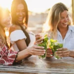 Multietniske piger jubler med cocktails i en bar på stranden - fokus på de forreste hænder, der holder drinksene