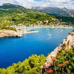 Panorama of Port Soller Mallorca with mountain peak Puig de la Bassa, marina Tramontana, beaches Platja de Can Generos and Platja des Traves from left to right with boulder and flowers in foreground.